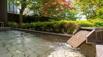 A tranquil outdoor water feature with a stone basin and a small waterfall, surrounded by lush greenery and trees.