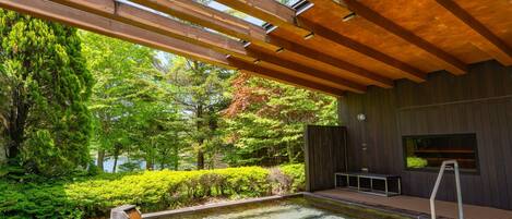 A modern outdoor pool area with wooden beams overhead, surrounded by lush greenery and a view of trees.