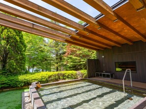 A modern outdoor pool area with wooden beams overhead, surrounded by lush greenery and a view of trees.