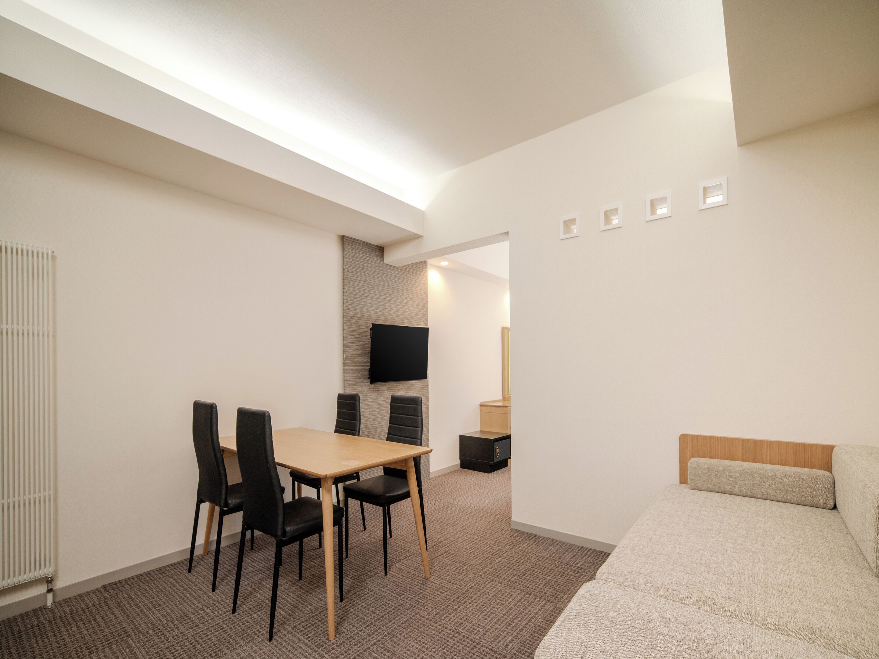 A modern hotel room with a wooden dining table, black chairs, a flat-screen TV mounted on a textured wall, a small wooden cabinet, and a beige sofa with a wooden headboard.