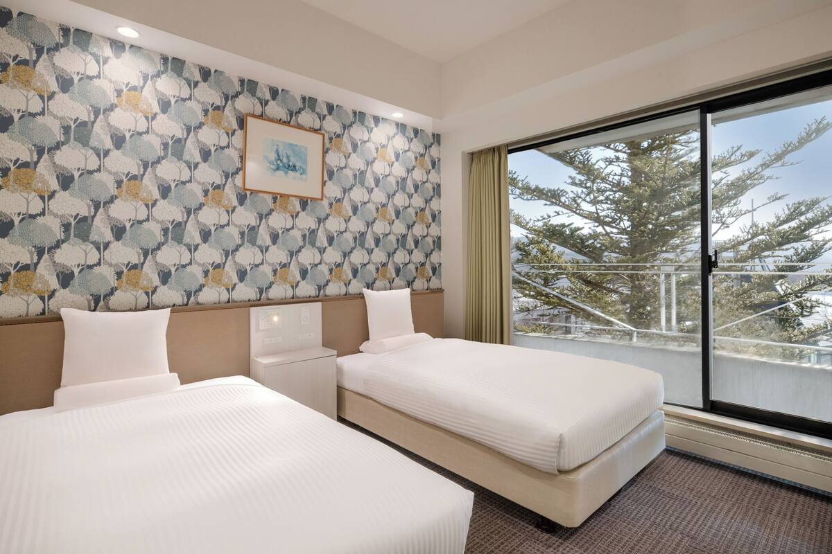 Ein Hotelzimmer mit zwei Betten, einem großen Fenster mit Blick auf Bäume, einer Wand mit ornamentalem Tapetenmuster und einem gerahmten Bild.