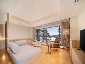 Ein modernes Wohnzimmer mit Sofa, einem kleinen Tisch, einem Sessel, einem Fernseher und einem großen Fenster mit Blick auf Bäume und ein Gewässer.
