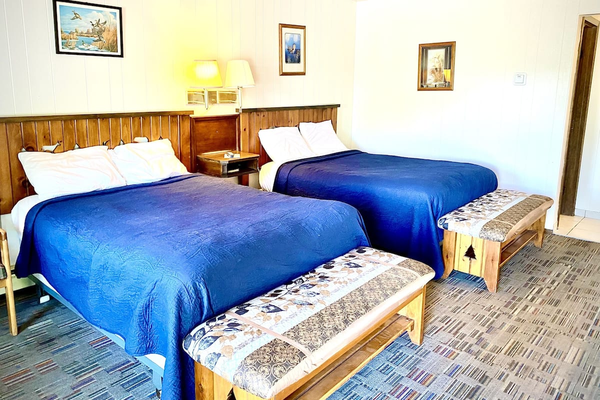 Quarto de hotel com duas camas, um banco, uma mesa de cabeceira e um abajur.