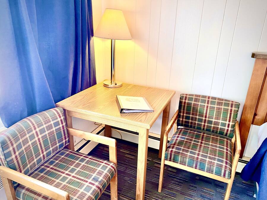 Quarto pequeno com uma mesa de madeira, duas cadeiras xadrez, um abajur e um livro sobre a mesa.