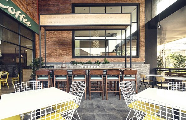 Um café ao ar livre com paredes de tijolo, grandes janelas e diversos tipos de assentos, incluindo mesas e cadeiras.