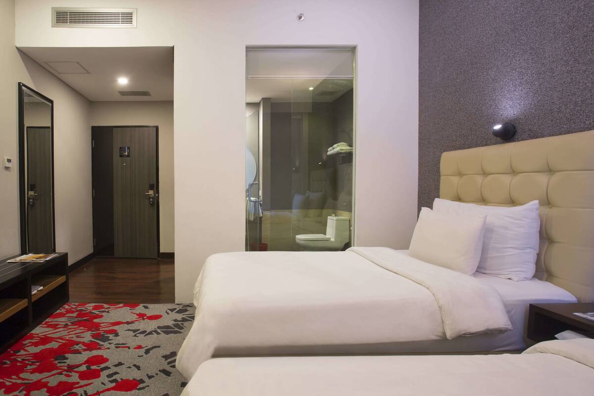 A hotel room with a bed, a nightstand, a mirror, a door, and a bathroom visible through a glass partition.