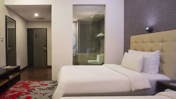 Habitación de hotel con cama, mesita de noche, espejo, puerta y baño visible a través de un panel de vidrio.