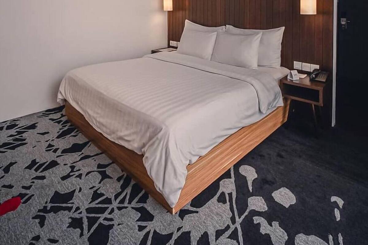 A modern hotel room with a wooden headboard, a large bed with white linens, two bedside lamps, and a nightstand with a phone and papers.