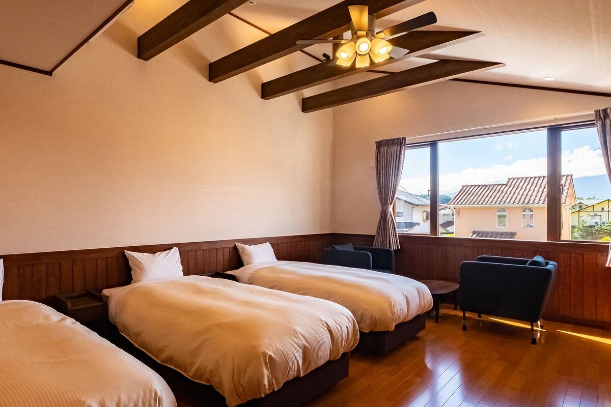 A hotel room with two beds, a wooden ceiling, a window with curtains, and a view of buildings outside.