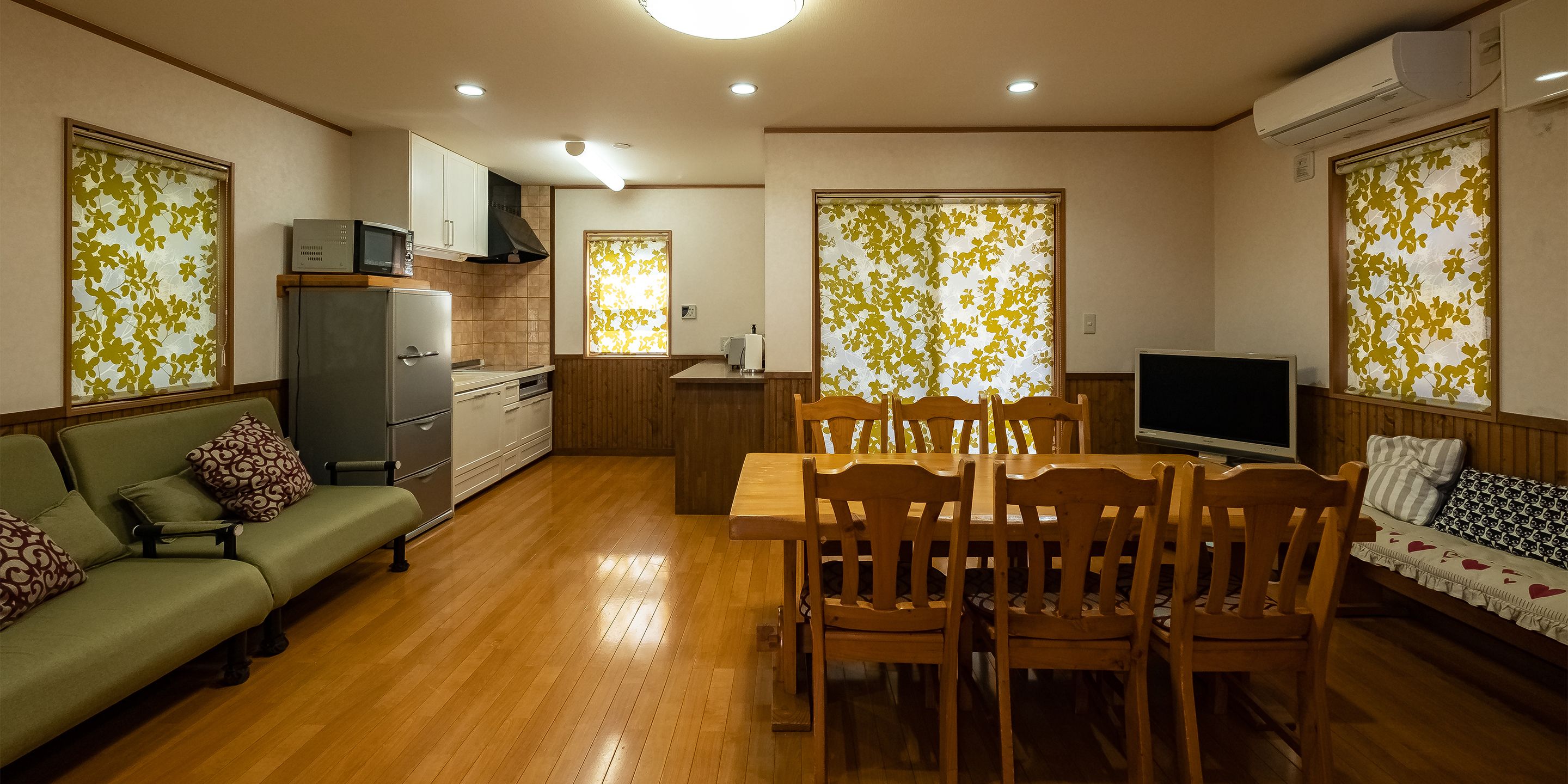 A compact living space with a dining table, wooden chairs, a sofa, a TV, and a kitchen area.