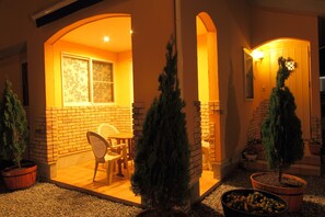 Uma área de pátio bem iluminada, com paredes de tijolo, uma mesa e cadeiras de madeira e árvores perenes em vasos.