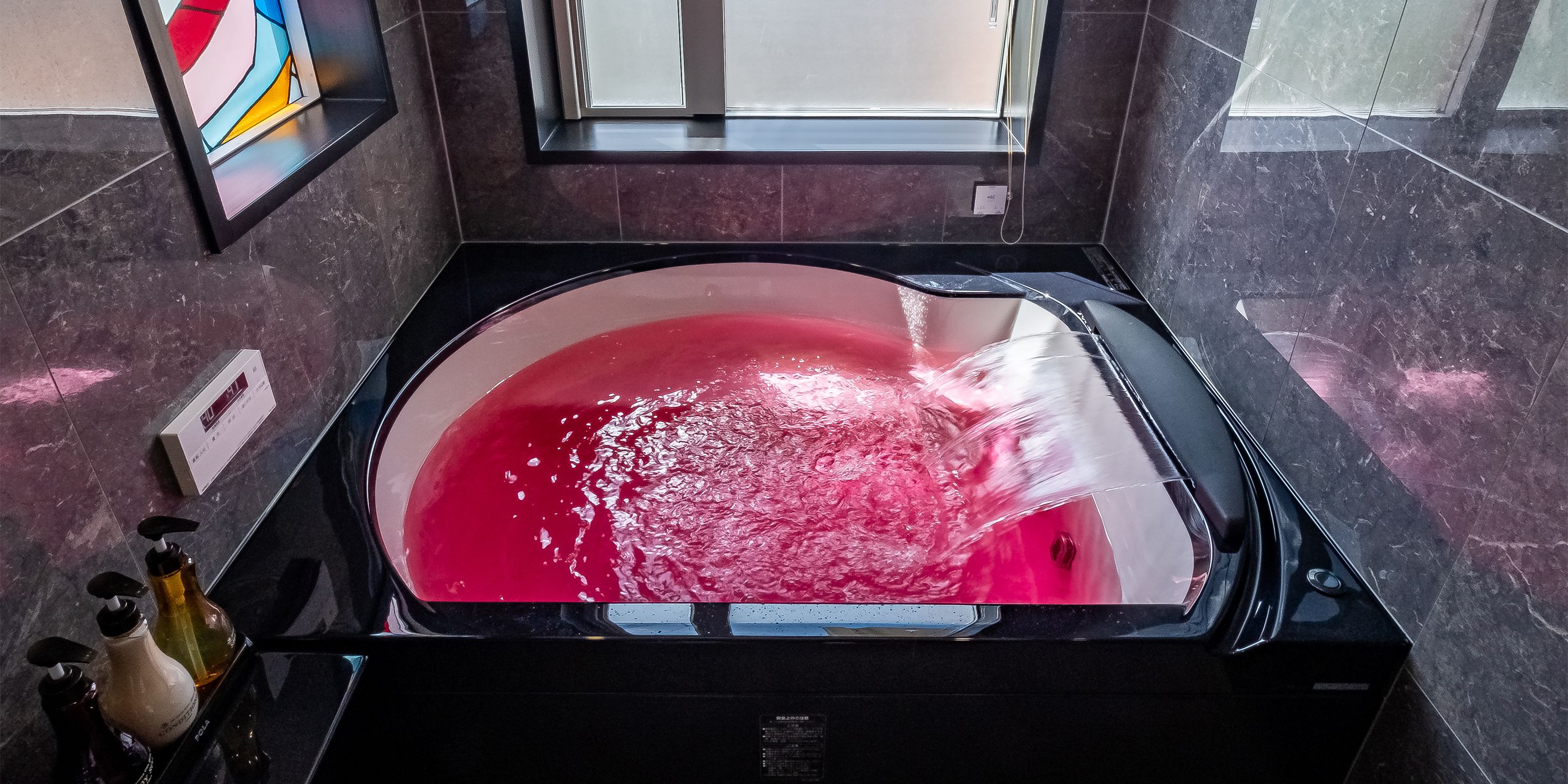 A modern bathroom with a large, black bathtub filled with red water, marble walls, and a window above the tub.