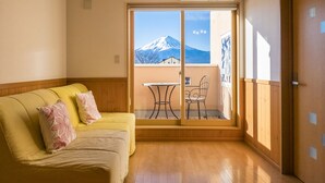 A room with a yellow sofa, a wooden floor, and a sliding glass door leading to a balcony with a view of a mountain. - Villa Ensoleille (Fujikawaguchiko)