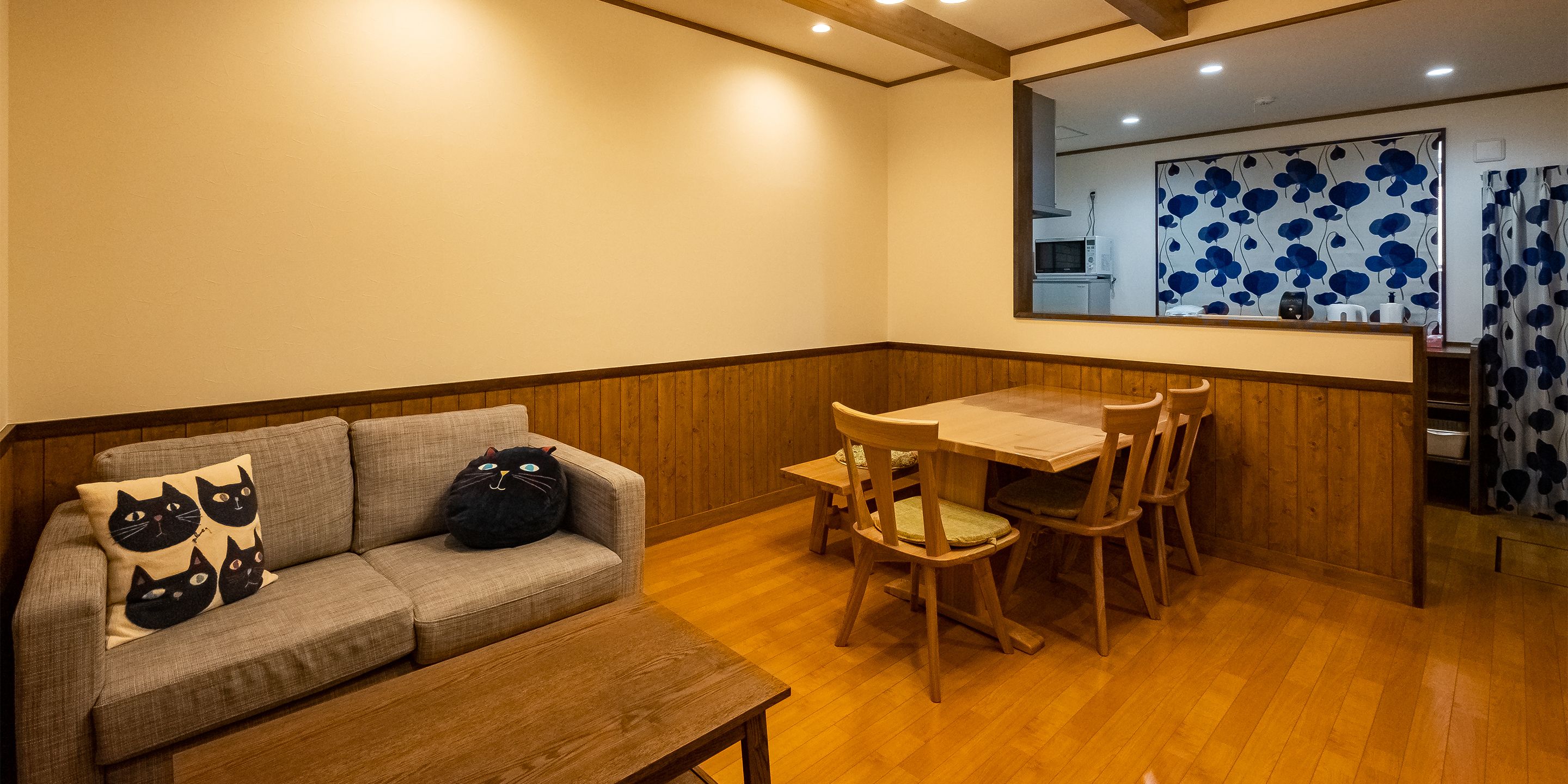 A cozy living area with a sofa, a wooden table, and chairs. A kitchen is visible through a window with a blue floral pattern.
