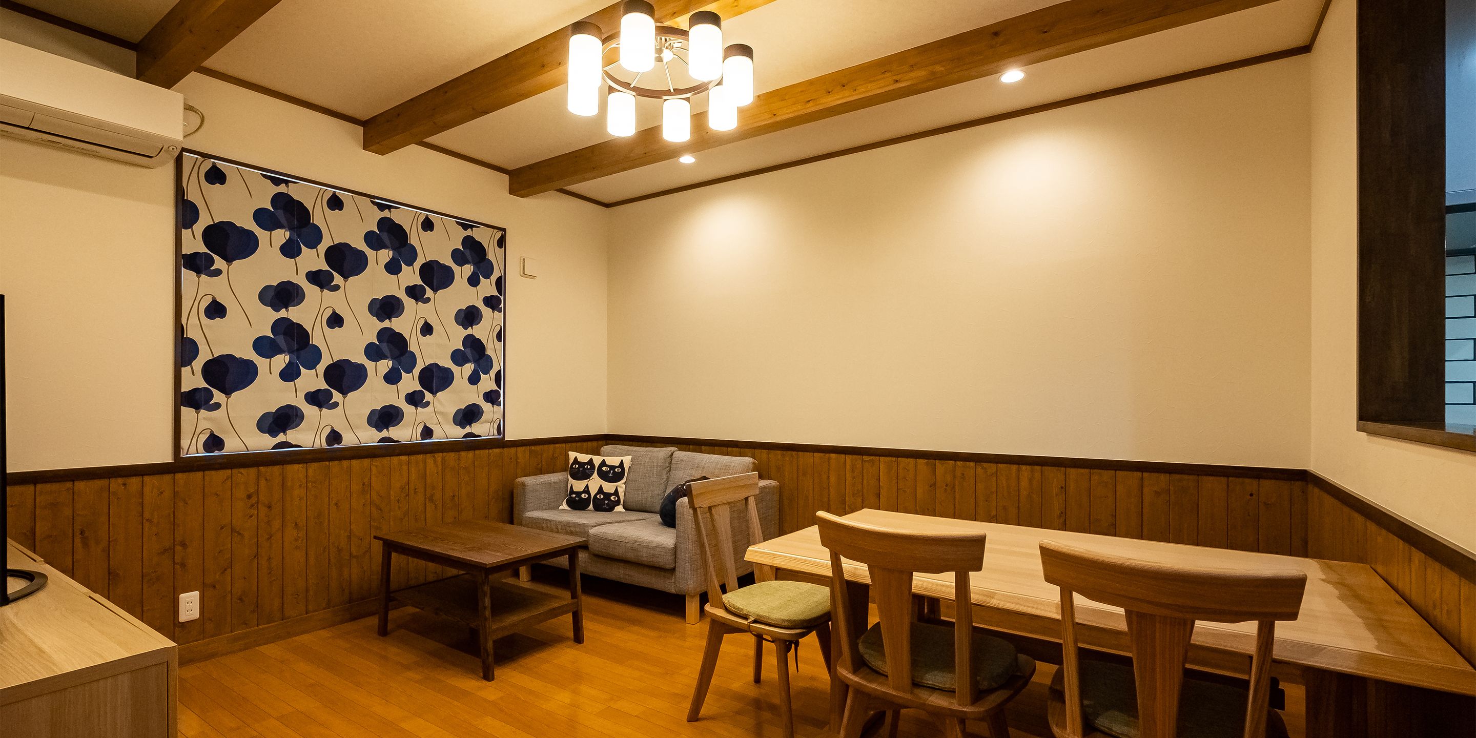 A cozy room with a wooden floor, a sofa with cat-themed cushions, a small table, and a dining table with chairs.