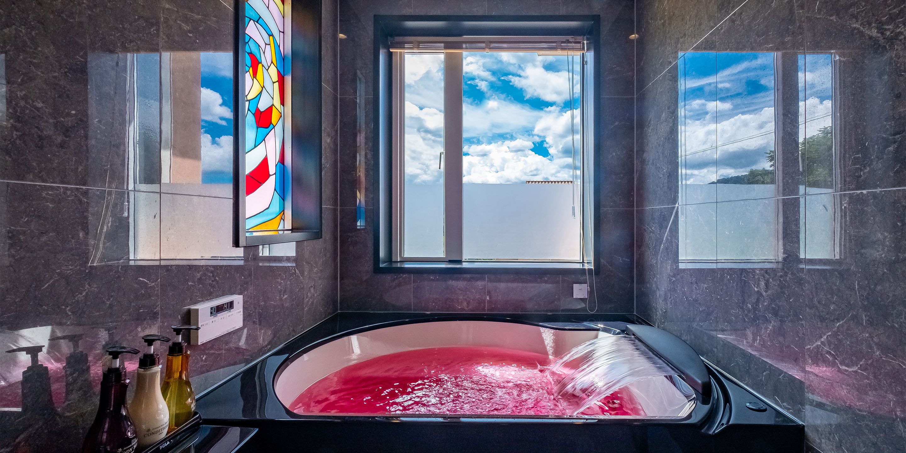 A modern bathroom with a large bathtub, a stained glass window, and a view of the sky and clouds.