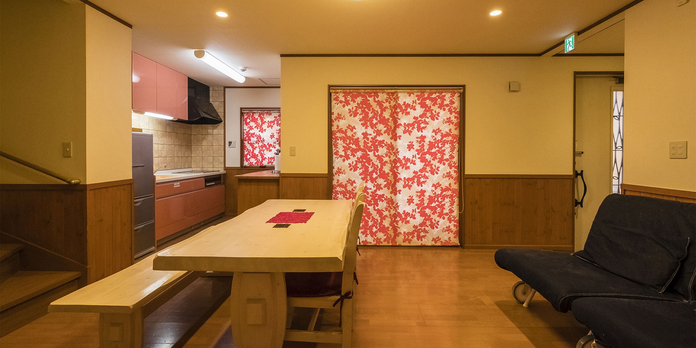 Un coin repas avec une table et des bancs en bois, un canapé noir et une porte rouge à motifs floraux.