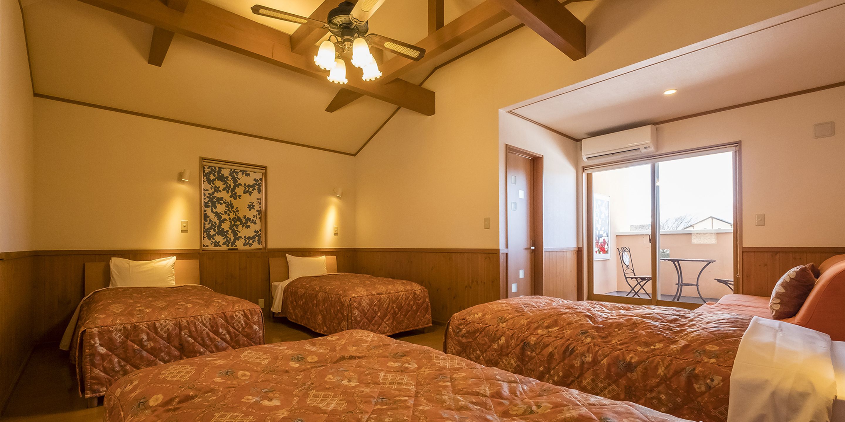 A hotel room with three beds, wooden beams on the ceiling, a ceiling fan, and a large window with a view of the outside.