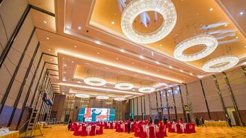 Un salón amplio con varias mesas redondas y sillas, un escenario con una pantalla grande, y un techo alto con candelabros circulares.
