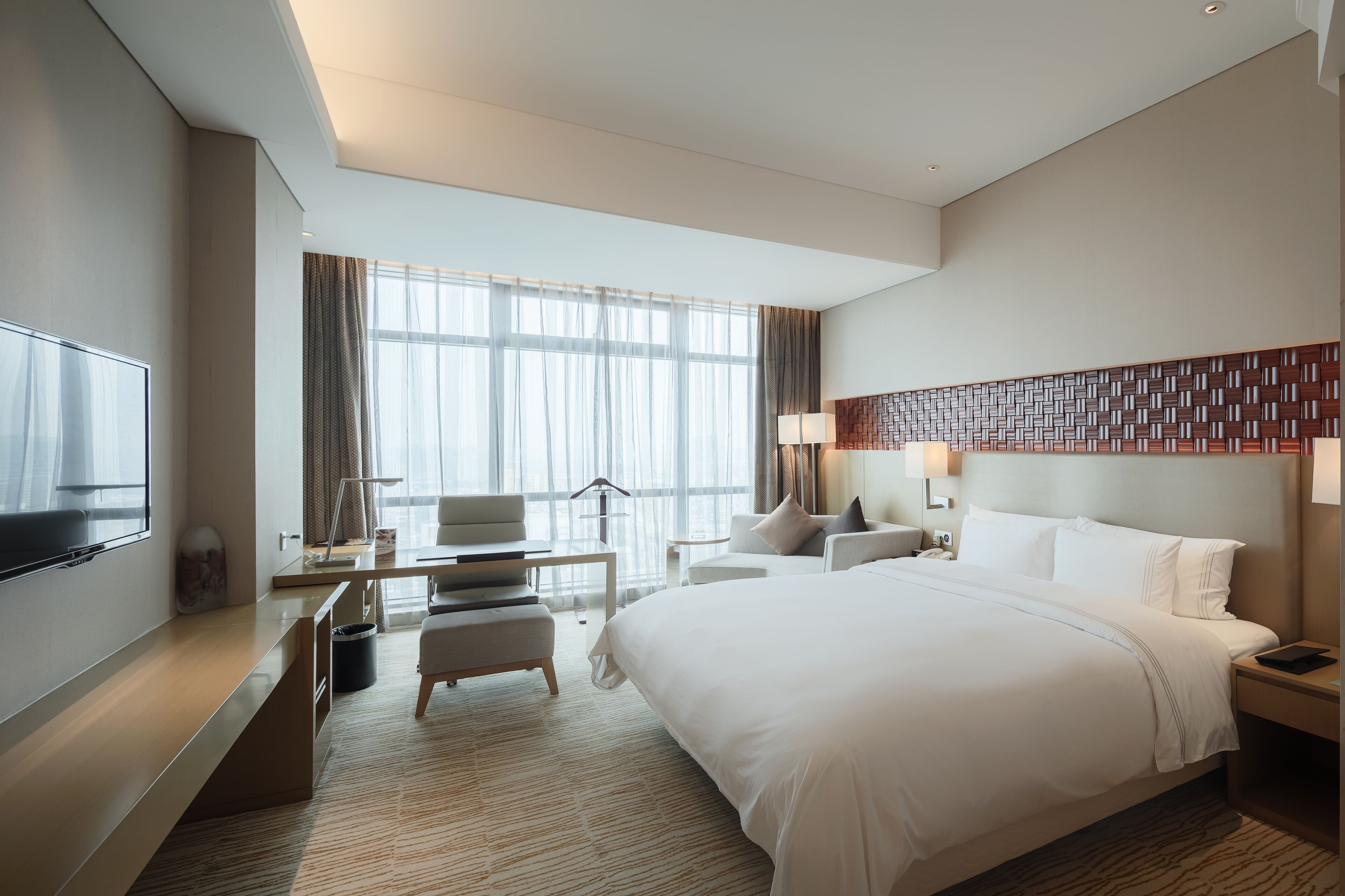 A modern hotel room with a large bed, a desk, a TV, and a view of the city.