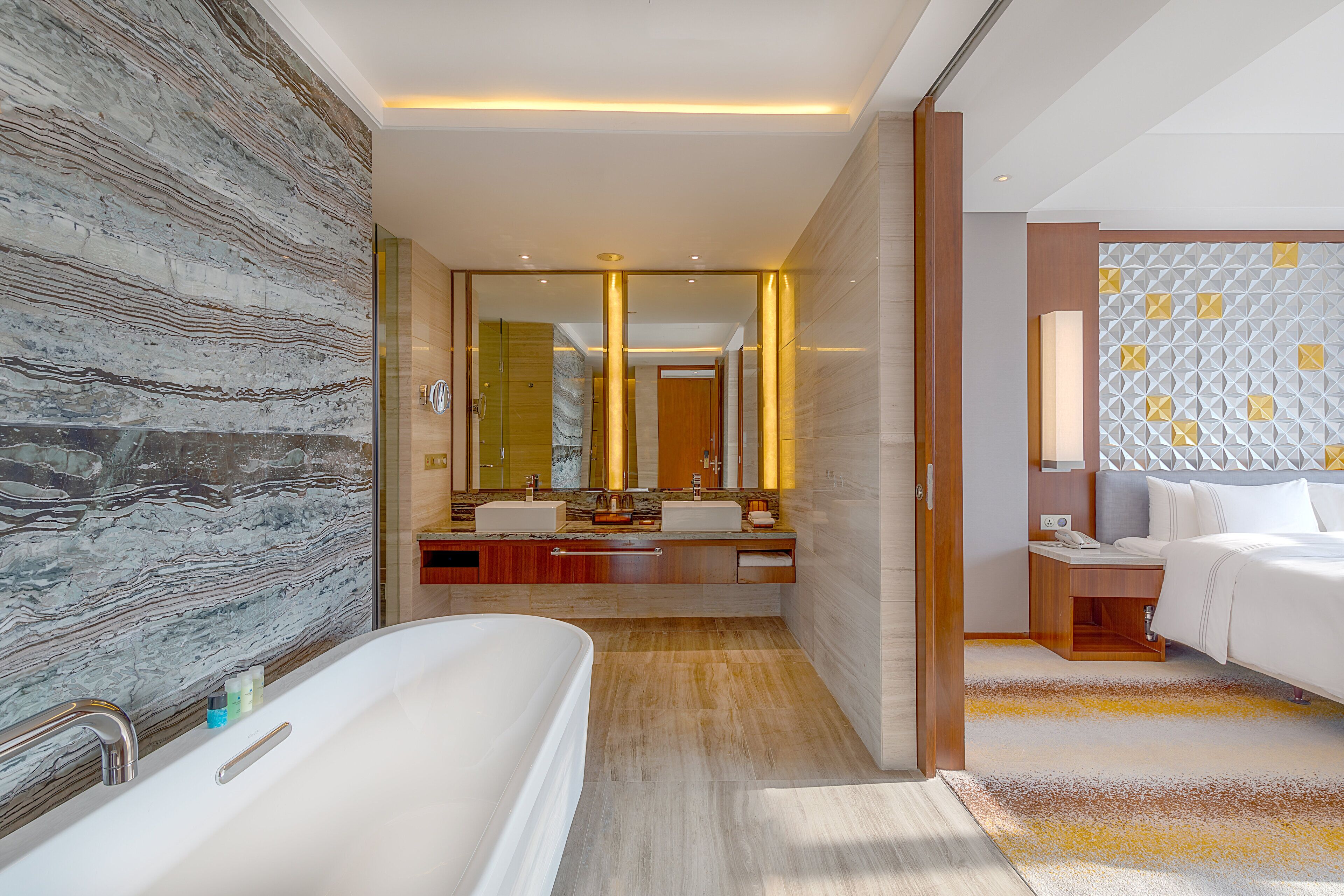 A modern bathroom with a marble wall, a large bathtub, a vanity with a mirror, and a bedside table with a lamp.