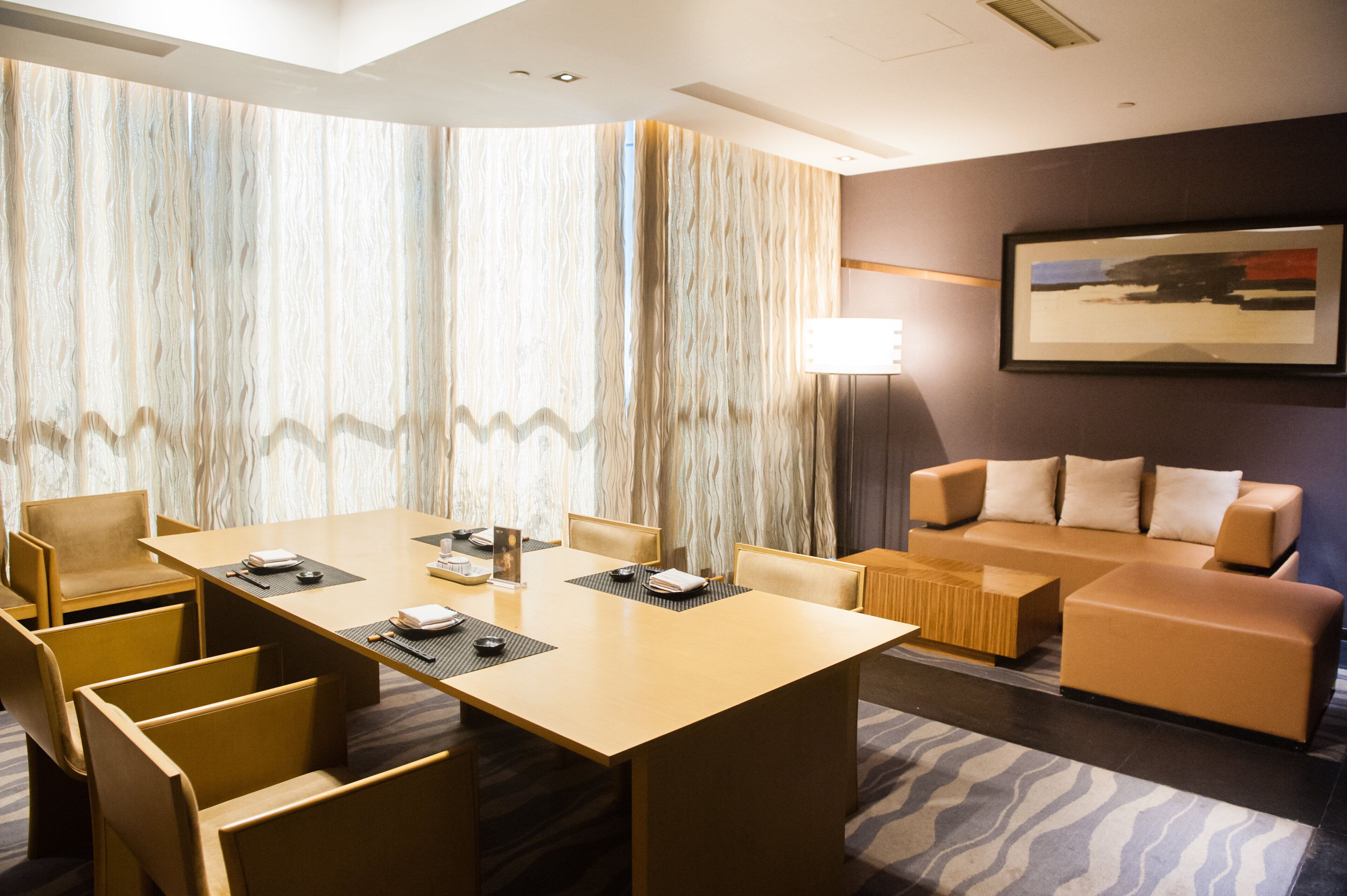 A modern hotel room with a large conference table, comfortable seating, a large window with curtains, and a framed picture on the wall.