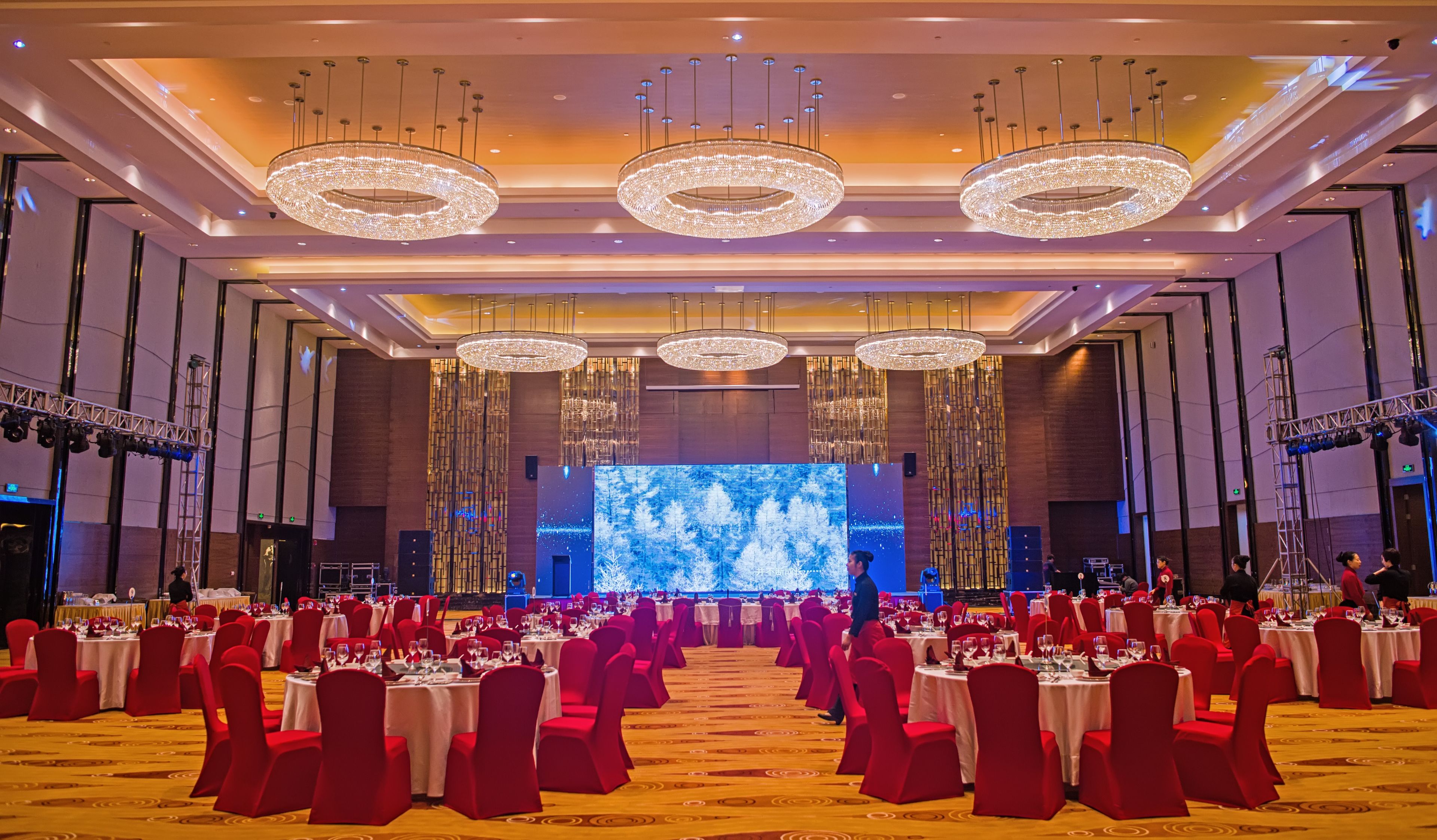 A large banquet hall with multiple round tables, red chairs, and chandeliers.