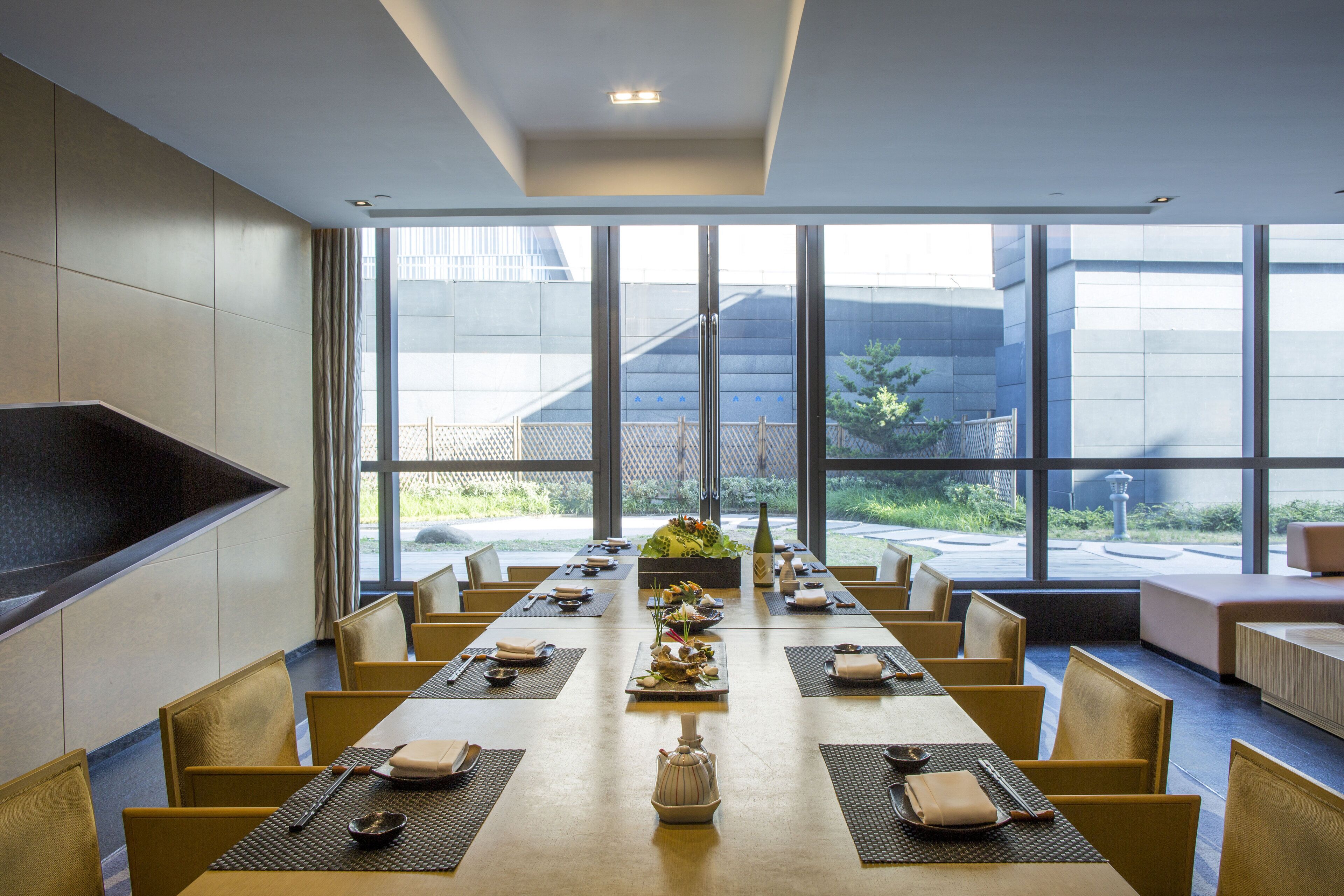 A modern dining area with a long table set for a meal, large windows, and a view of the outdoors.