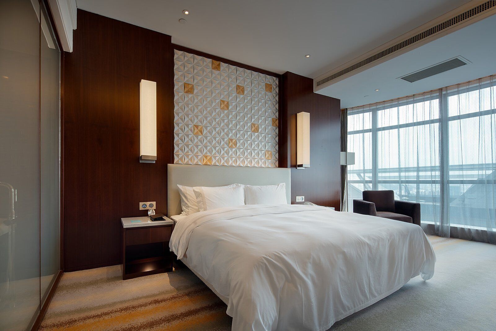 Una habitación de hotel moderna con una cama grande, una mesita de noche, una silla y un ventanal con vistas a la ciudad.