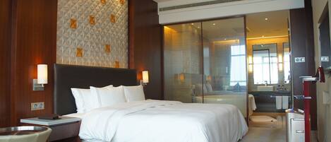 Une chambre d’hôtel moderne avec un grand lit, une cloison en verre, une salle de bain avec un miroir et un porte-serviettes rouge.
