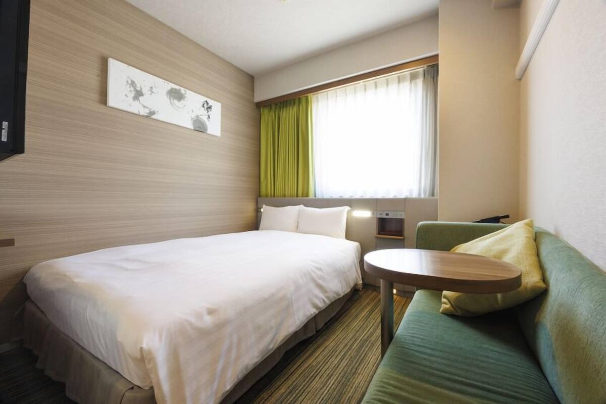 A hotel room with a bed, a green sofa, a small table, a window with curtains, and a wall-mounted artwork.