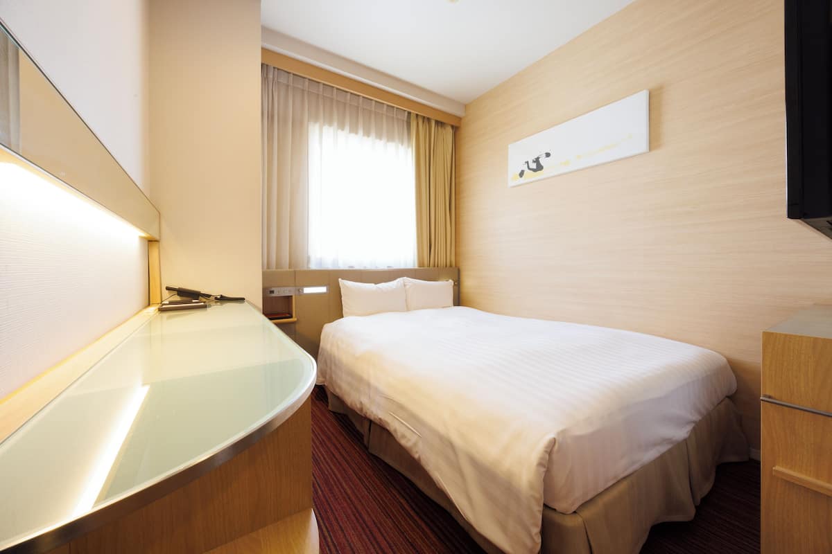 A hotel room with a bed, a bedside table, a mirror, a window with curtains, and a wall-mounted artwork.