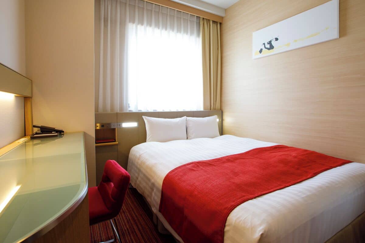 A hotel room with a bed, a desk, a chair, a window with curtains, and a wall-mounted artwork.