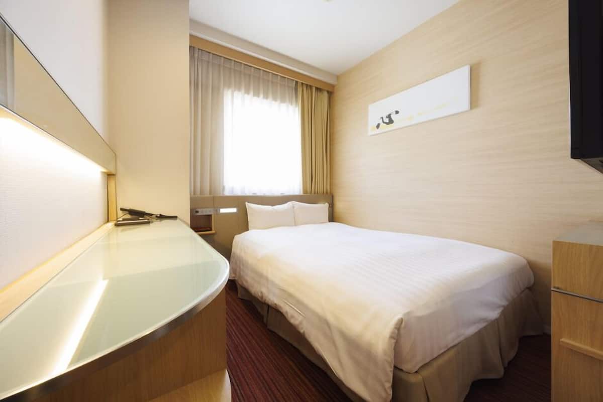A hotel room with a bed, a bedside table, a mirror, a window with curtains, and a wall-mounted artwork.