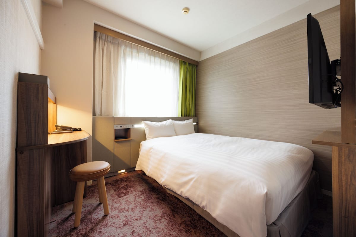 A hotel room with a bed, a desk with a chair, a TV, and a window with curtains.