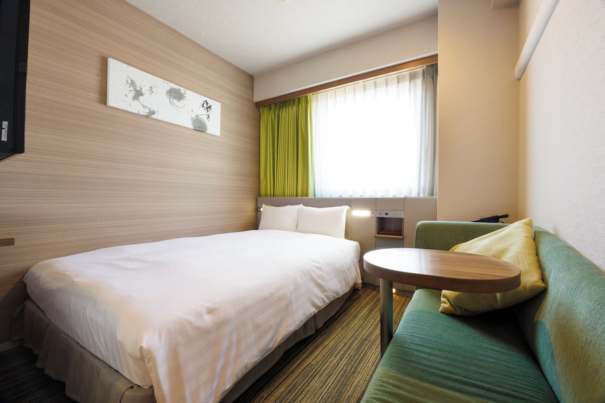 A hotel room with a bed, a green sofa, a small table, and a window with curtains.