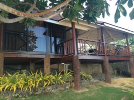 Uma casa de madeira de dois andares com varanda, rodeada por plantas tropicais e um muro de pedra.