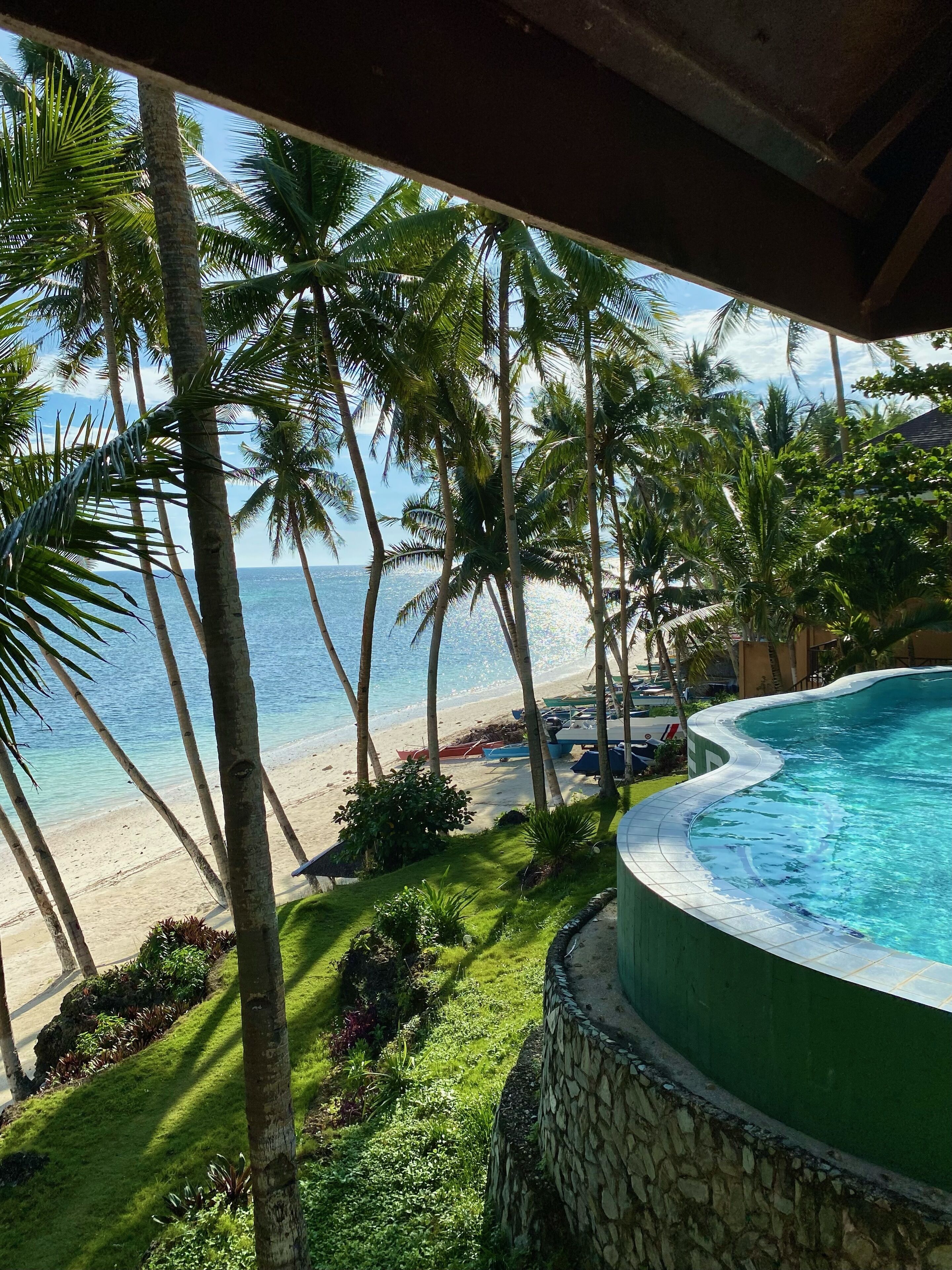 A pool with a stone wall, surrounded by palm trees and a beach view.
