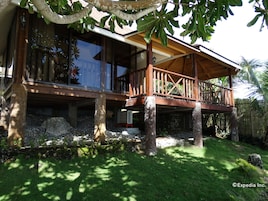 A two-story wooden house with a balcony, surrounded by lush greenery and a stone wall.