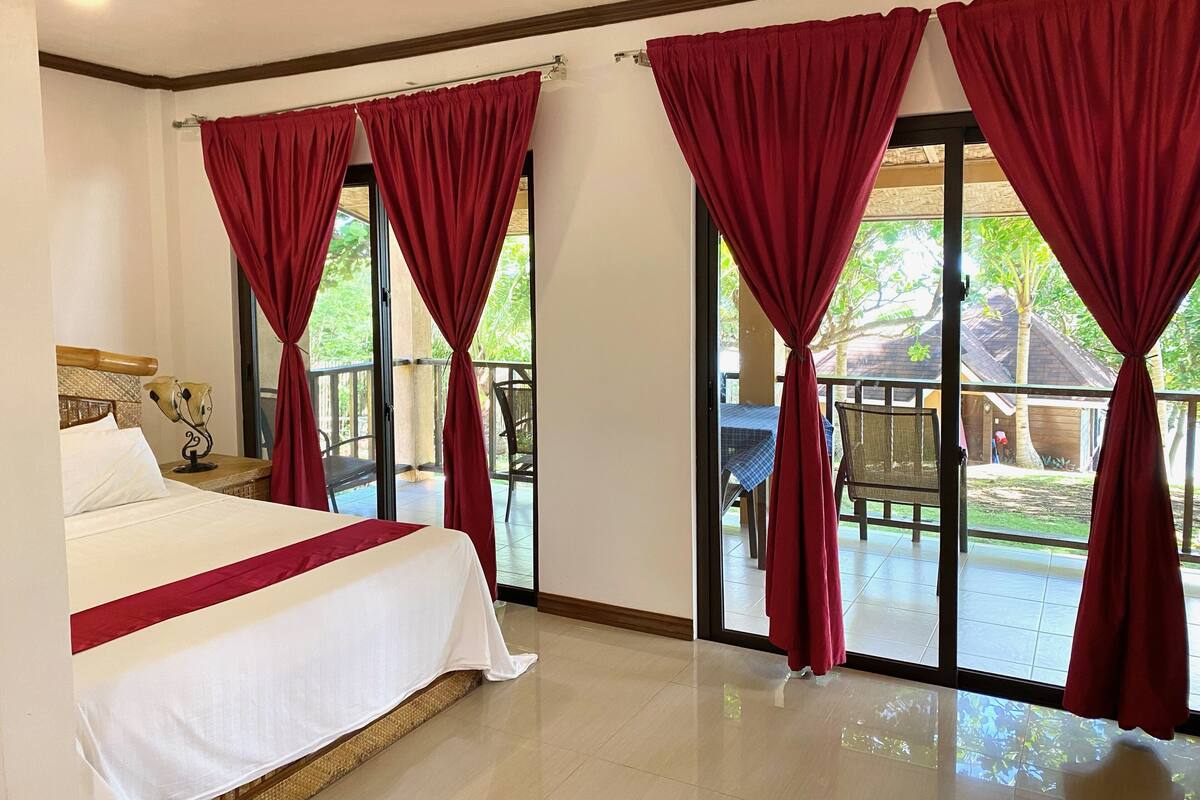 A hotel room with a bed, a nightstand, a lamp, and a view of the outdoor area through large windows with red curtains.