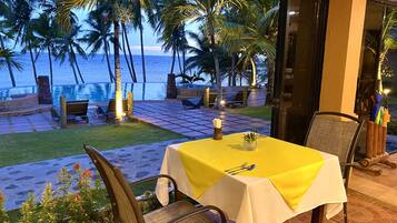 Una mesa para dos, con mantel amarillo, con vista a una playa con palmeras y una piscina.