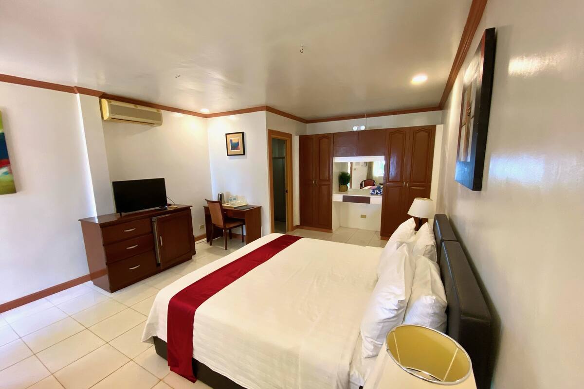 A hotel room with a large bed, a desk with a chair, a television, and a kitchenette area.