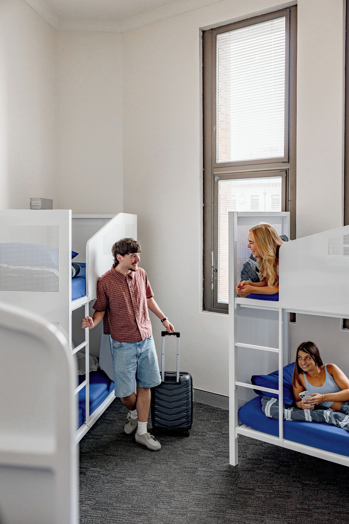 A dormitory room with bunk beds, a window with blinds, and a suitcase on the floor.