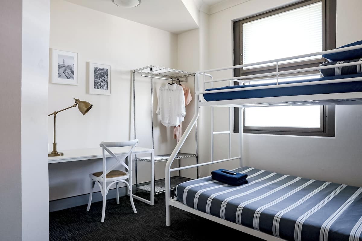 Una cama individual superpuesta con escritorio y silla, un perchero con ropa y una ventana con persianas.