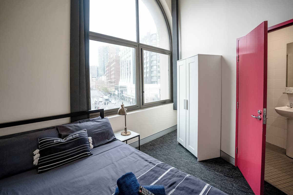 Habitación de hotel con un ventanal grande, cama con ropa de cama gris, lámpara de noche, armario y baño con puerta roja.