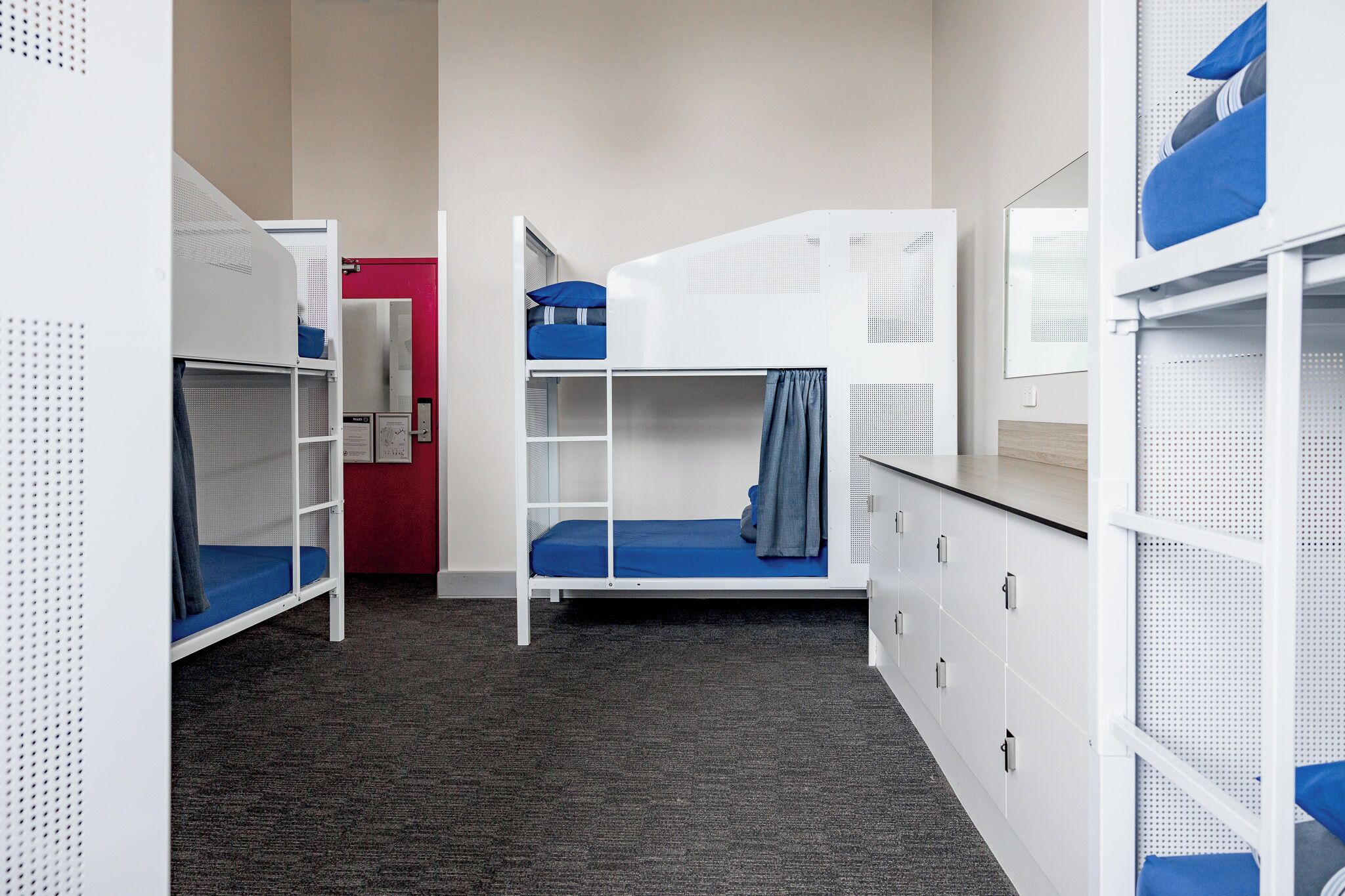A dormitory room with bunk beds, a red door, and a white wardrobe.
