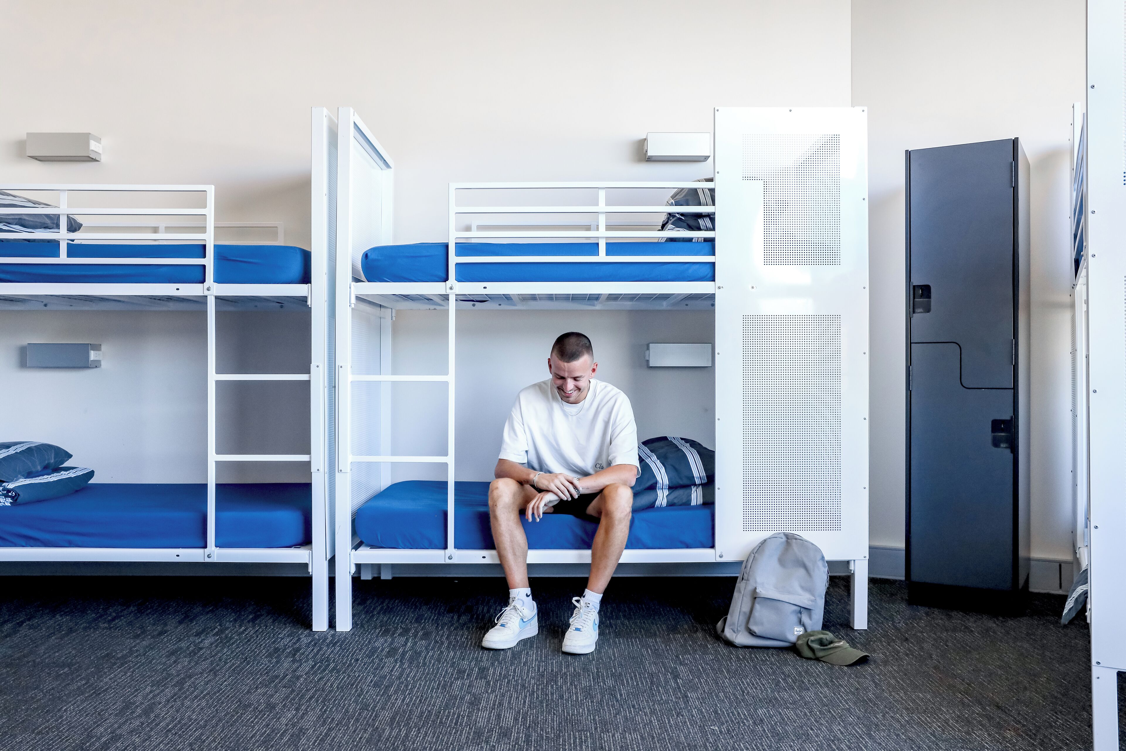 A bunk bed room with a person sitting on the lower bunk, a grey suitcase, and a door to the right.