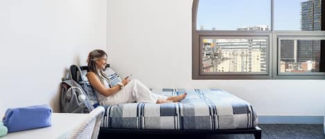 Una persona sentada en una cama en una habitación con un ventanal grande, un escritorio y una silla.