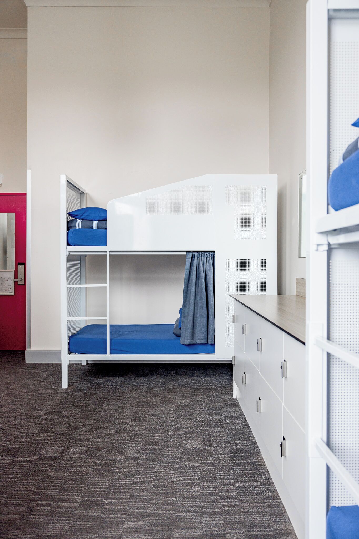 A bunk bed room with a desk and drawers, carpeted floor, and a door with a red frame.