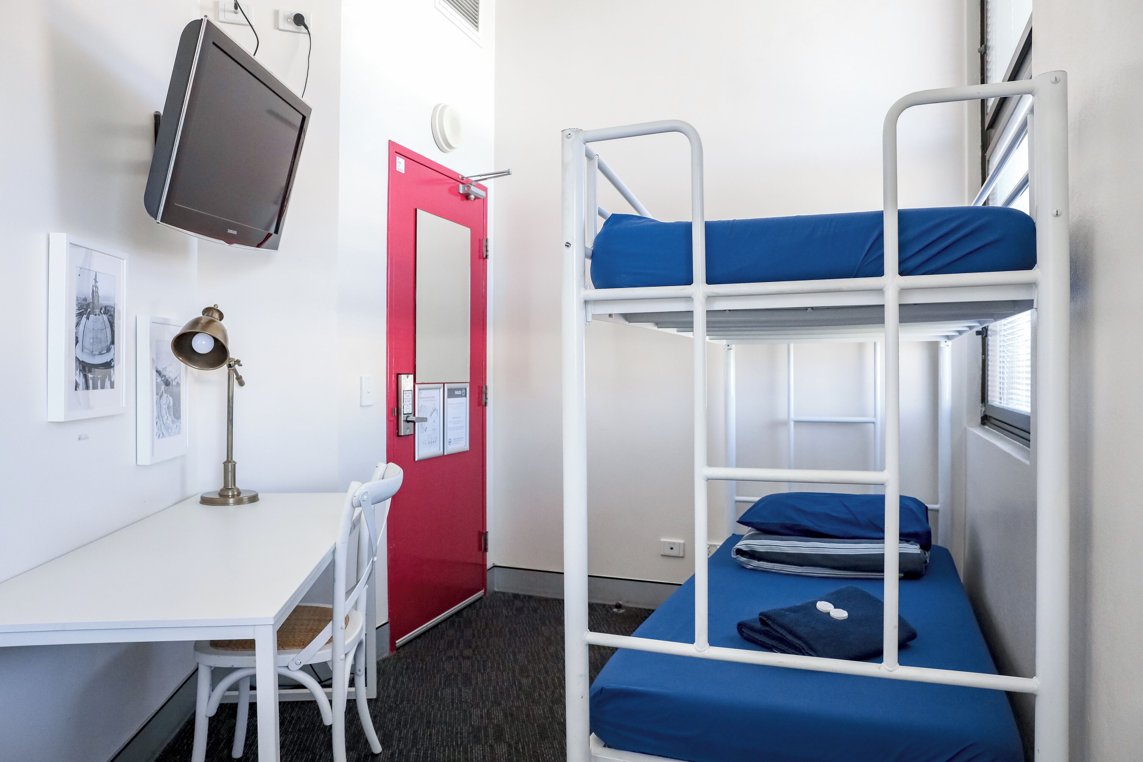 A bunk bed with blue bedding, a desk with a chair, a red door, and a mounted television.